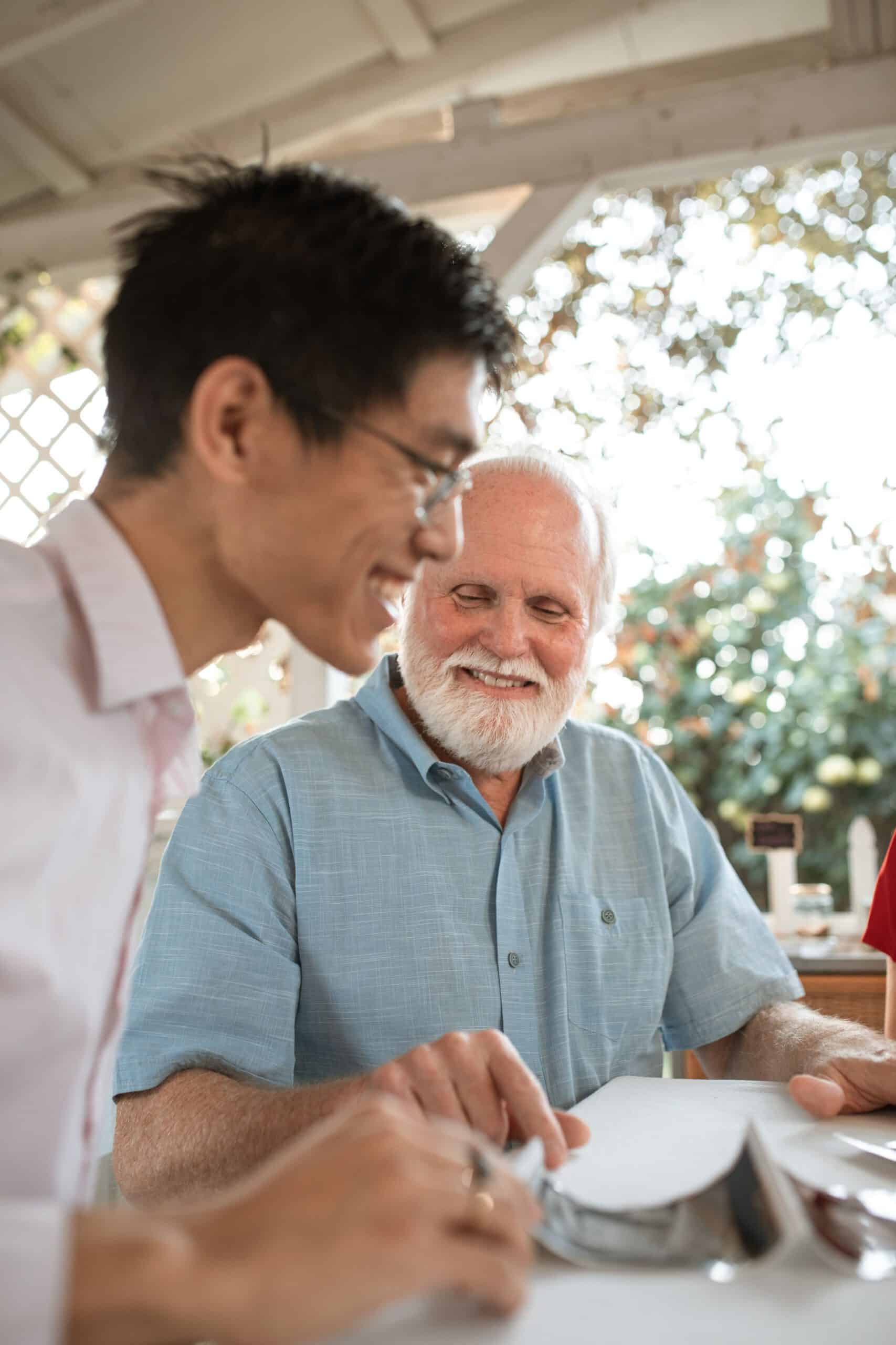 Two men of different generations — one younger, one older — seated together at a table, heads close, sharing a warm and unguarded moment of conversation. The older man smiles with quiet joy. A soft-focus gathering is visible in the background.